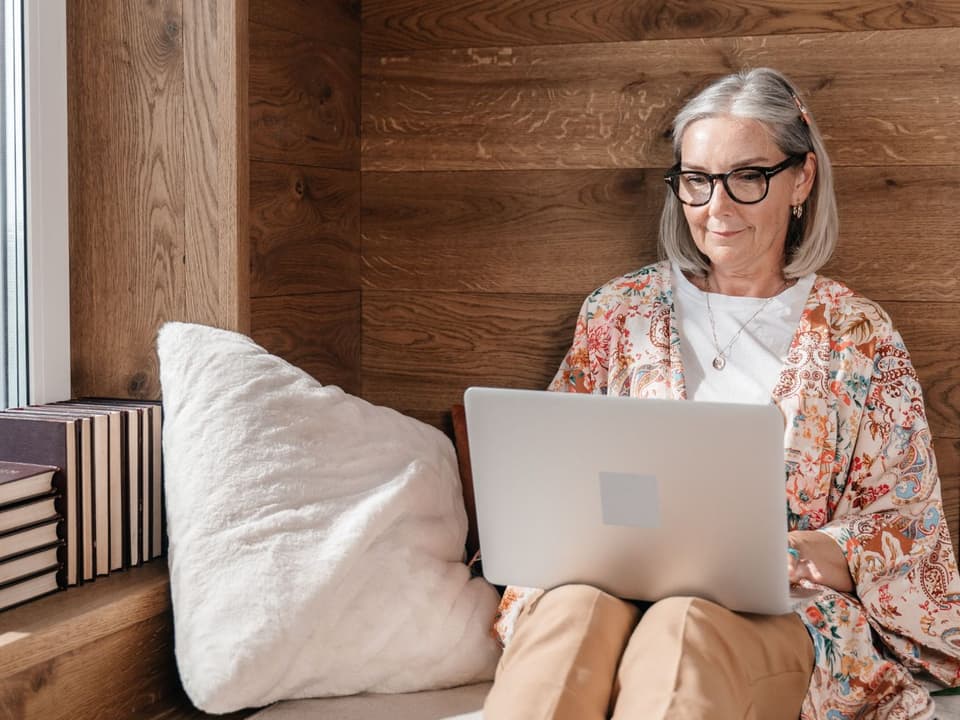 An older woman in glasses reading on her laptop while relaxing on a window seat at home