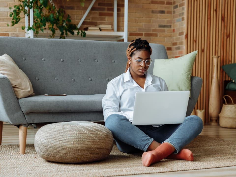 A woman sitting cross-legged on her living-room floor, typing on a laptop